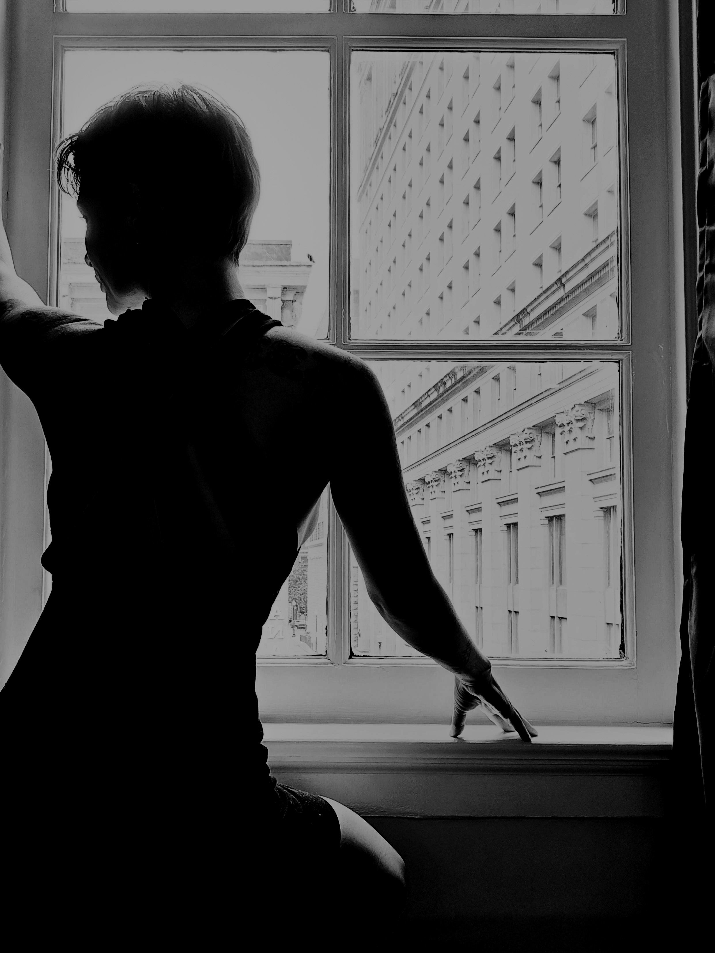 Heather seated in profile at a tall window, seen from behind, tattoos across her shoulders. Through the panes, a stone building across the street. Black and white.